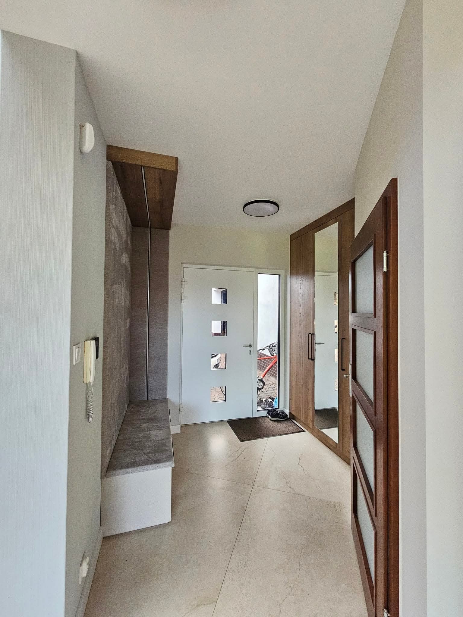 Wykończony korytarz z szafą z lustrem i siedziskiem w Linowcu. Nowoczesny design, jasne kolory, minimalistyczne detale. Widok na drzwi wejściowe i podłogę z płytek.