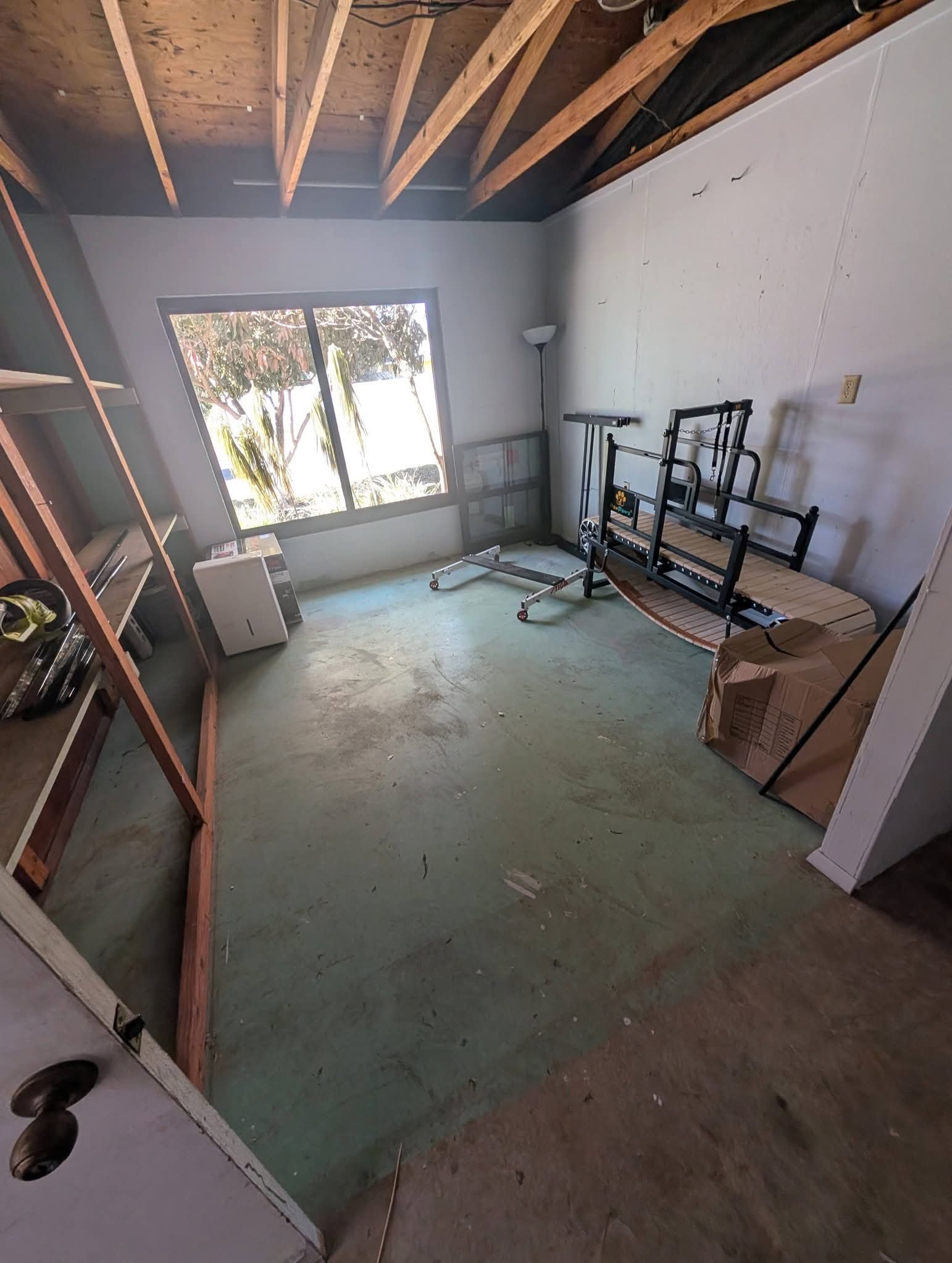 Pusta przestrzeń z oknem, częściowo umeblowana, z widoczną konstrukcją dachu i sprzętem do ćwiczeń. Podłoga wymaga sprzątania, widoczne ślady użytkowania.