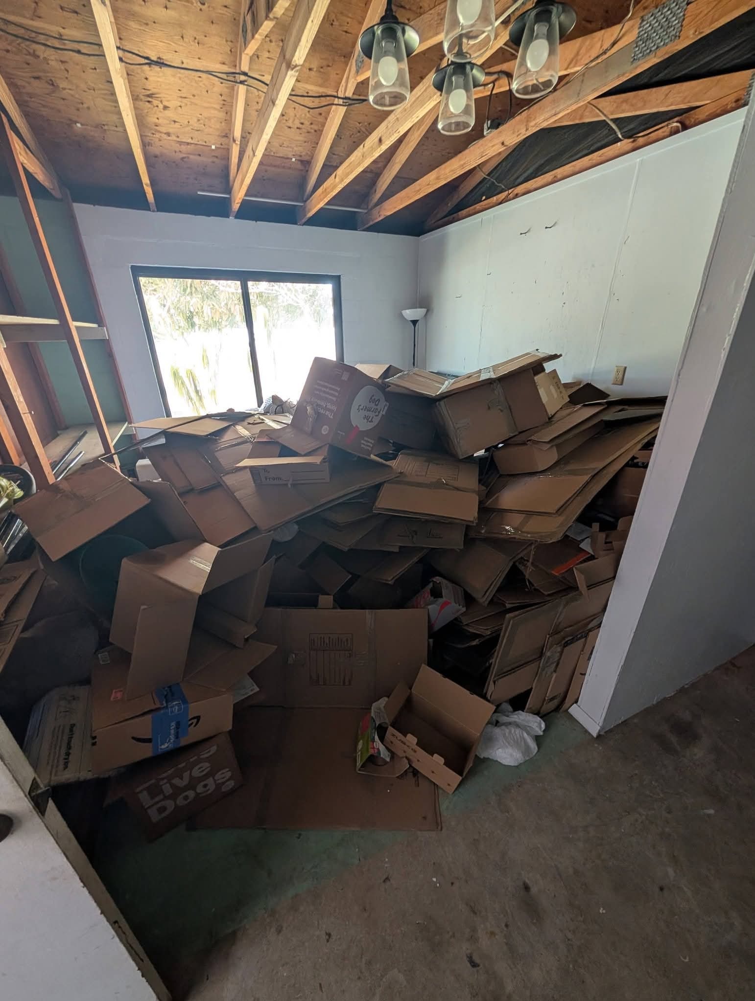 Pokój zagracony stertą kartonowych pudeł, widoczne okno i surowe wykończenie wnętrza. Chaos przed opróżnianiem mieszkania. Pudełka różnych rozmiarów.