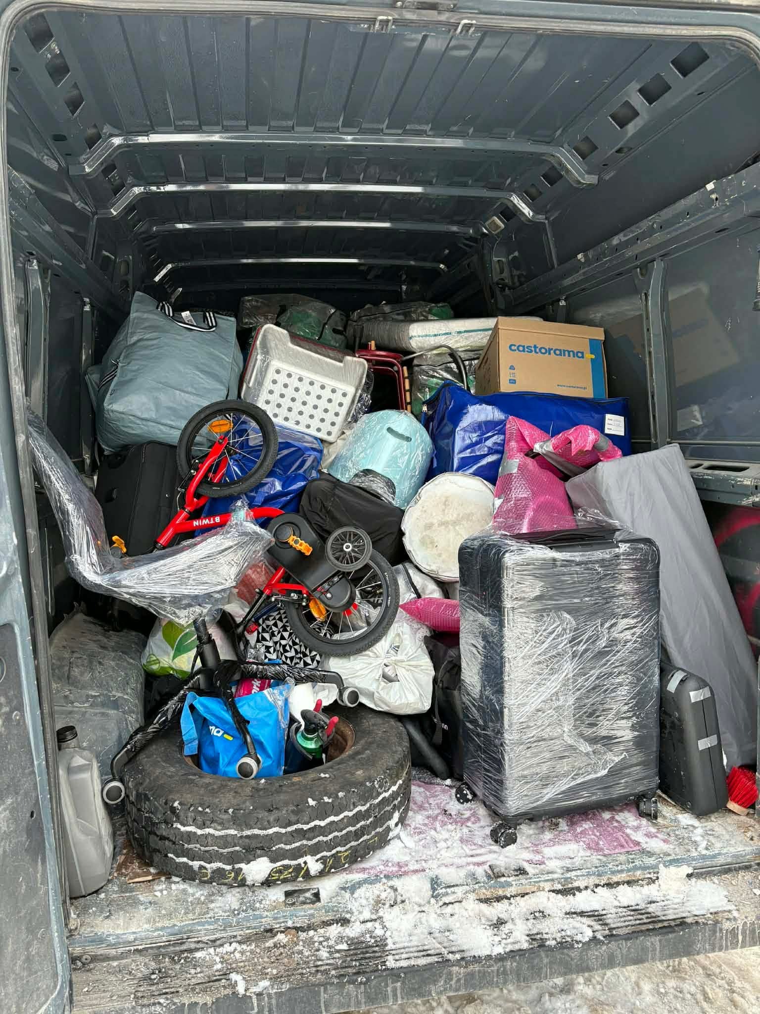 Wnętrze busa wypełnione po brzegi różnorodnymi przedmiotami, w tym rowerami dziecięcymi, walizkami owiniętymi folią, torbami i kartonem Castorama. Widoczny brud na podłodze.