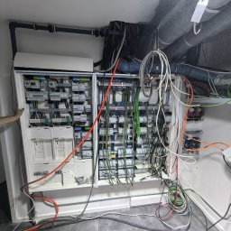 Pod-napięciem - Rozbudowana skrzynka elektryczna z licznymi przewodami w Szczecinie. Widoczne zabezpieczenia, bezpieczniki i kable różnej grubości. Częściowo otwarte drzwi szafki.