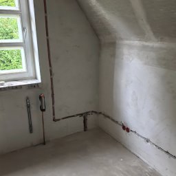 Pod-napięciem - Surowe wnętrze poddasza z instalacją elektryczną i rurami miedzianymi przy oknie. Widoczne narzędzia i puszki elektryczne w ścianie. Ściany i podłoga w stanie surowym.