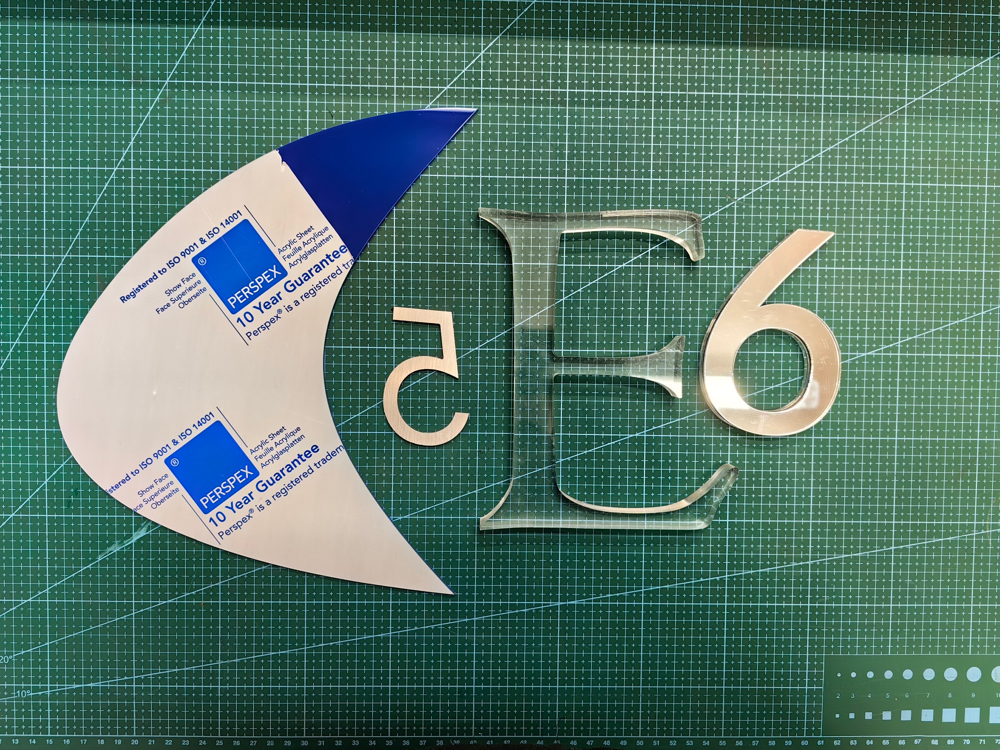 Płyta plexi po frezowaniu CNC: litera E z przezroczystego akrylu, cyfry 5 i 6 z metalizowanego tworzywa, szablon Perspex na zielonej macie pomiarowej.
