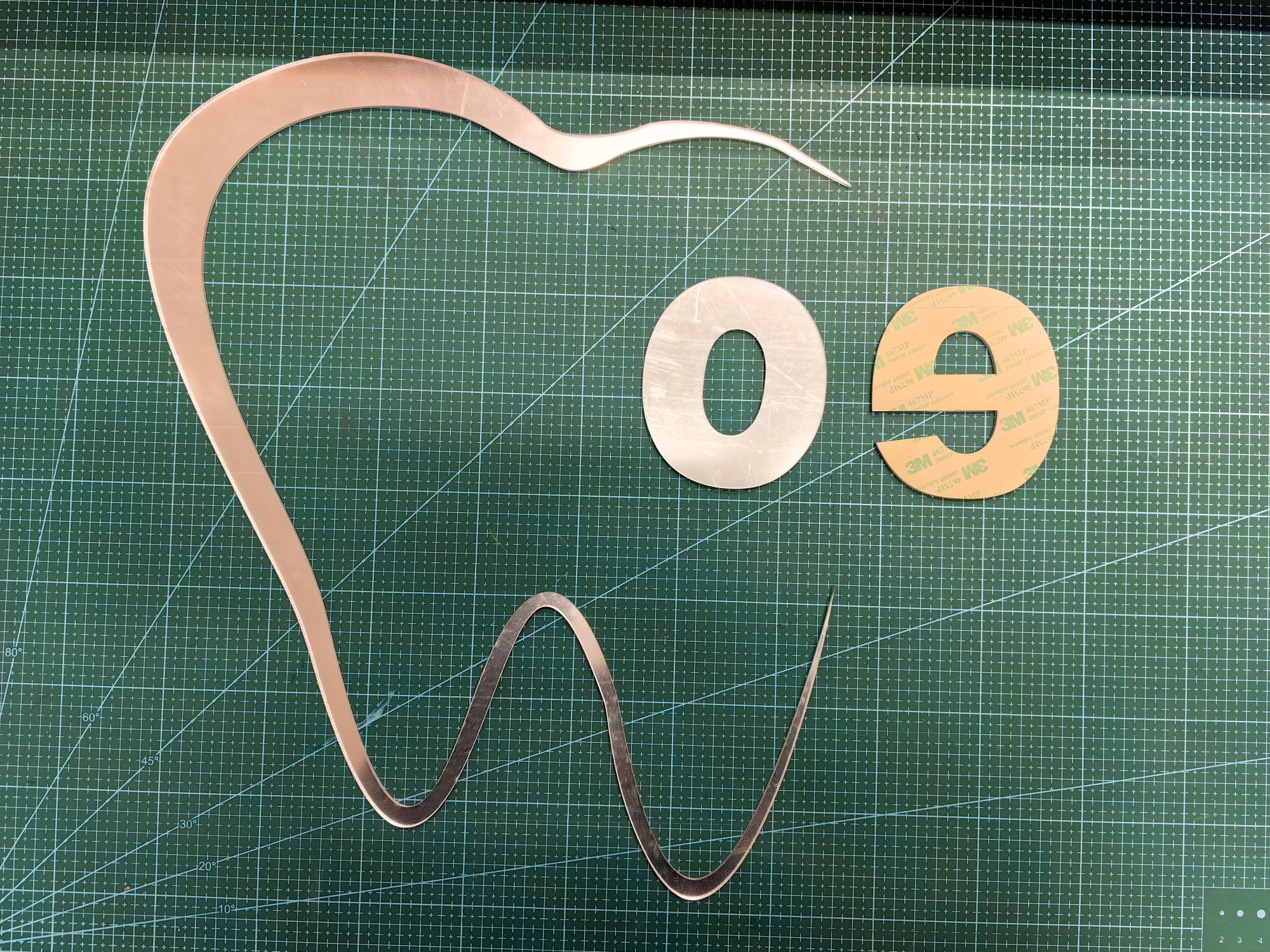 Elementy z plexi wycięte na ploterze: kształt zęba, litera 'O' i 'e' z taśmą 3M, leżące na zielonej macie do cięcia. Precyzyjne wycinanie kształtów.