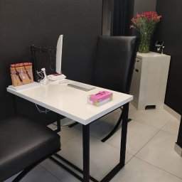 Trychologia Bednarek - Gabinet trychologiczny: białe biurko z modelem skóry głowy, monitor, klawiaturą i pudełkiem chusteczek. W tle czarna ściana i kwiaty w wazonie.