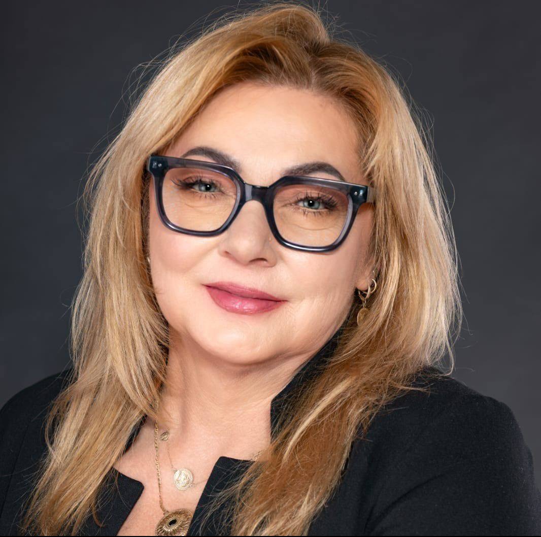 Portret kobiety w okularach i czarnej marynarce na ciemnoszarym tle. Widoczne złote kolczyki i naszyjniki. Uśmiech na twarzy, blond włosy.