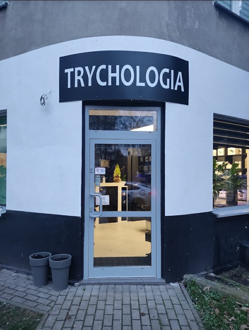 Wejście do gabinetu trychologicznego z szyldem 'Trychologia' nad drzwiami. Widoczne donice z roślinami przed wejściem i wnętrze gabinetu przez szklane drzwi.