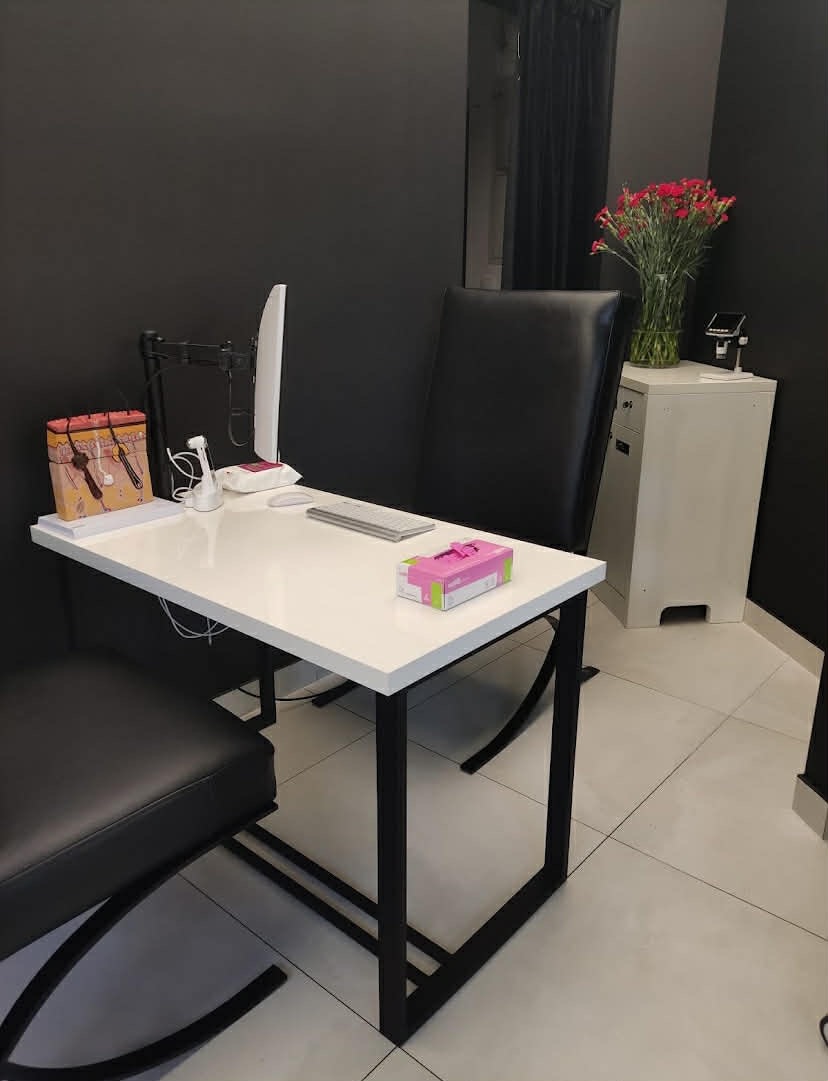 Gabinet trychologiczny: białe biurko z modelem skóry głowy, monitor, klawiaturą i pudełkiem chusteczek. W tle czarna ściana i kwiaty w wazonie.