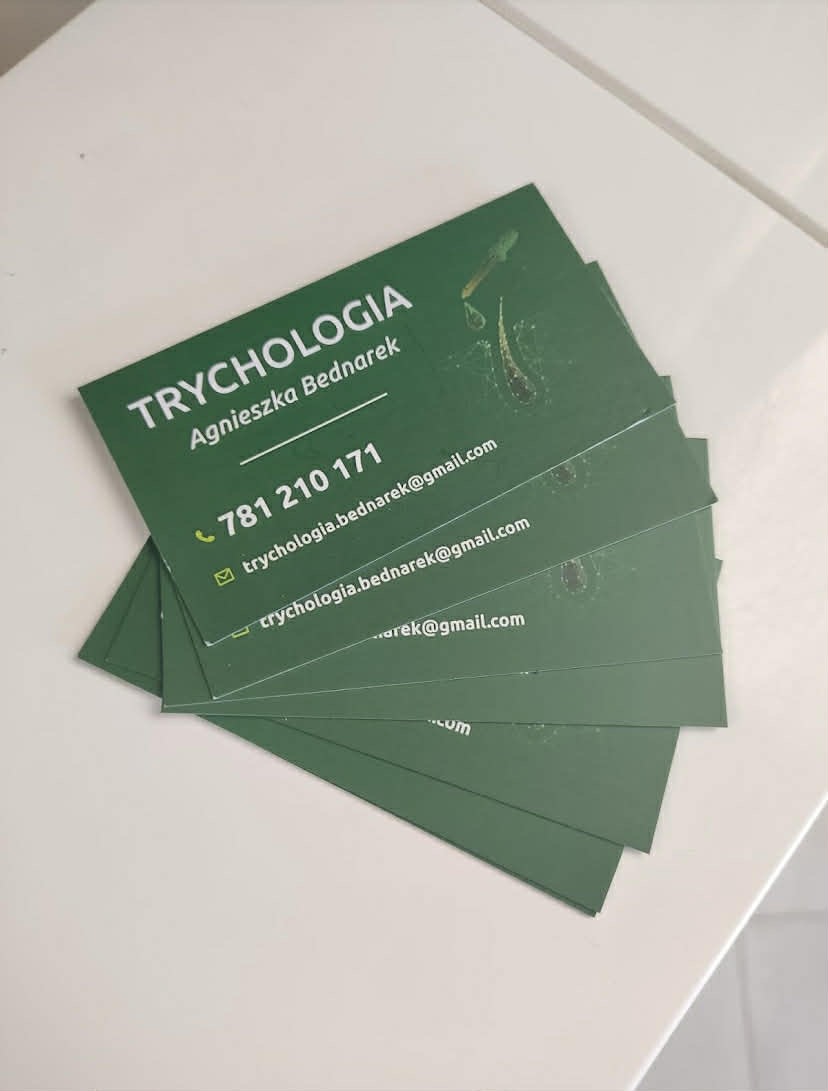 Stos zielonych wizytówek trychologicznych z danymi kontaktowymi, leżących na białym blacie. Widoczne logo z motywem włosów i pipety. Minimalistyczny design.