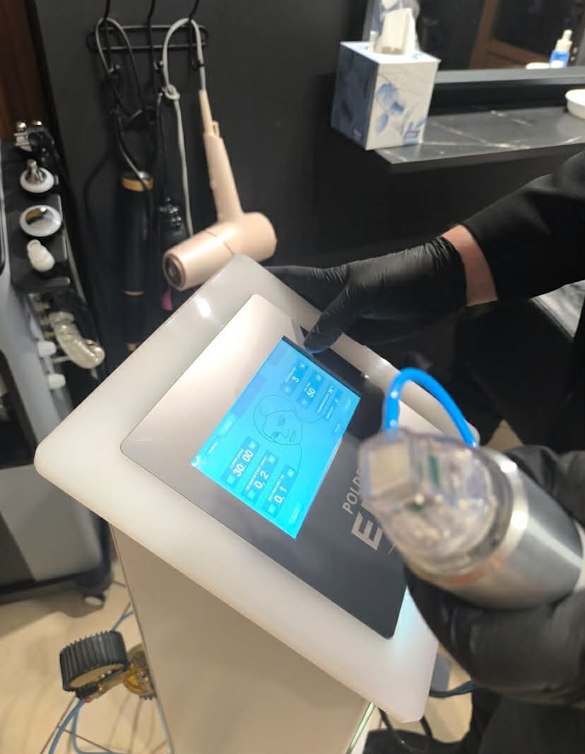 Trycholog w czarnych rękawicach obsługuje urządzenie do zabiegów na skórę głowy z niebieskim ekranem dotykowym w gabinecie trychologicznym w Łodzi.