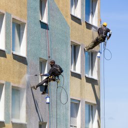 CleanPoint Group - Dwóch alpinistów przemysłowych czyści elewację budynku z użyciem myjek ciśnieniowych, zawieszeni na linach na tle błękitnego nieba.