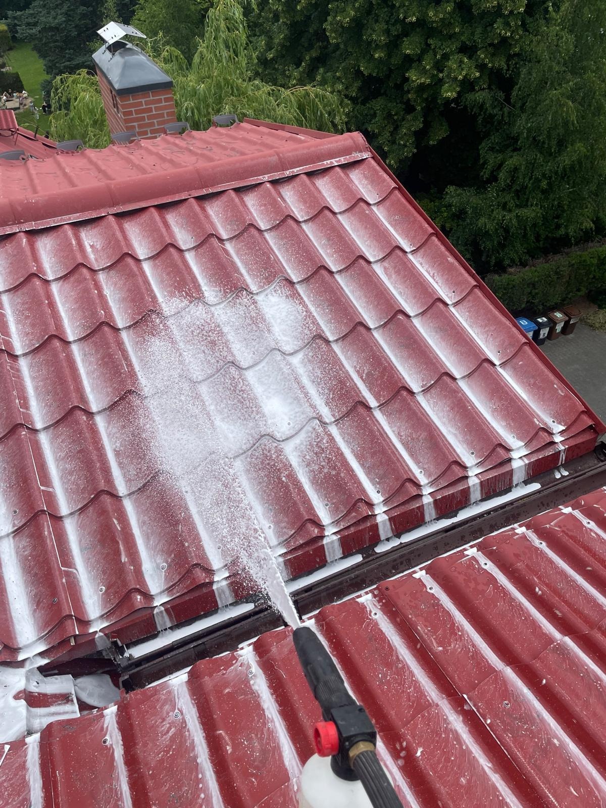 Czerwona dachówka czyszczona pod ciśnieniem z użyciem piany, widok z góry, fragment dachu po lewej stronie już umyty, w tle zieleń drzew.