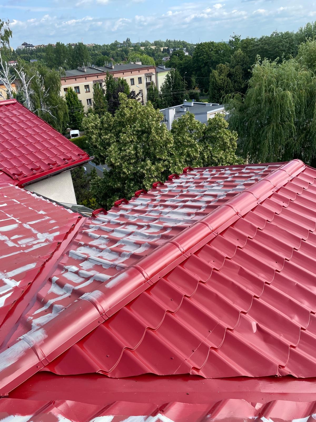 Częściowo pomalowany na czerwono dach z blachy trapezowej, widok z góry na tle zabudowy miejskiej i zieleni. Etap malowania dachu.