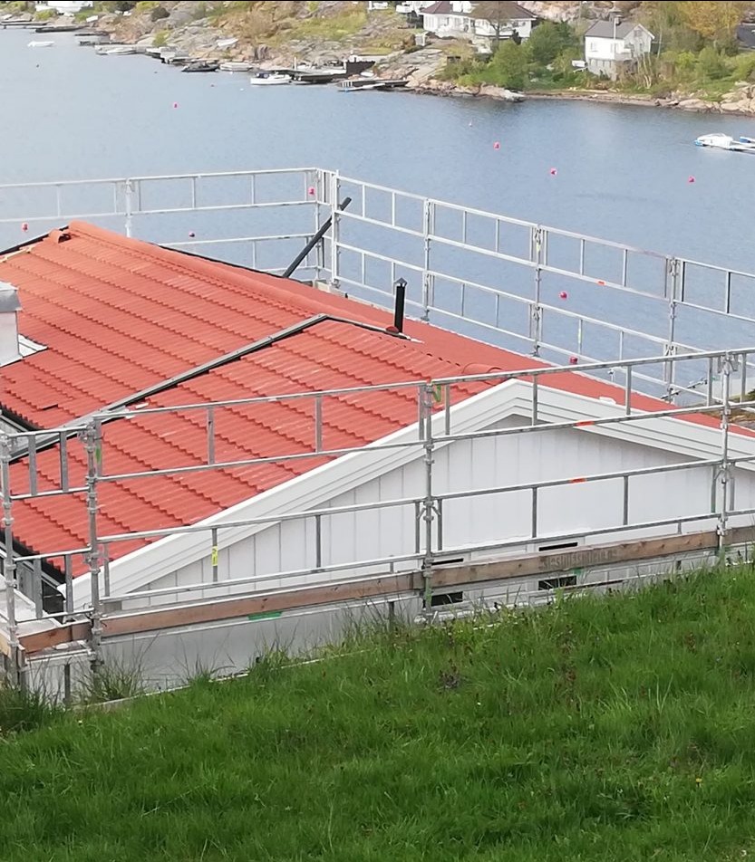 Dom z nową, czerwoną dachówką, otoczony rusztowaniem na tle jeziora i zielonego wzgórza. Widok z góry.