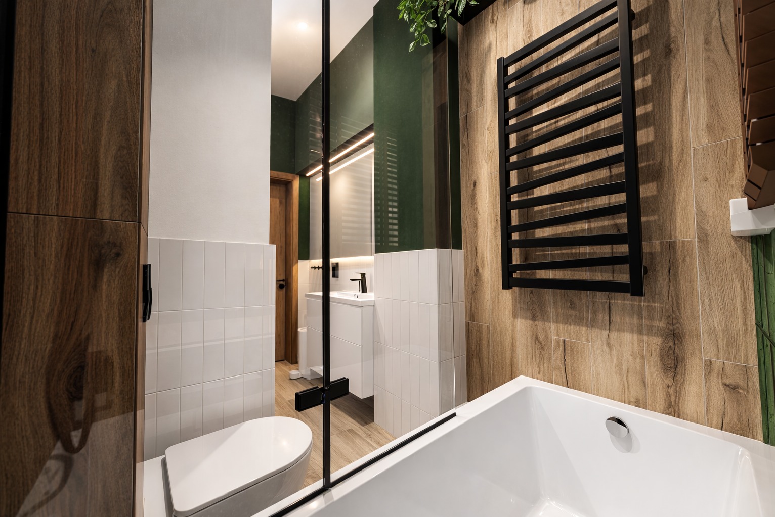 Nowoczesna łazienka z drewnopodobnymi płytkami, białą armaturą i czarnym grzejnikiem drabinkowym. Widoczne lustro z podświetleniem i zielony akcent kolorystyczny.