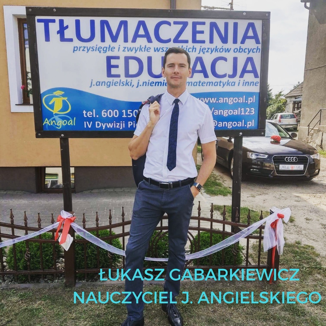 Mężczyzna w koszuli i krawacie stoi przed tablicą reklamową z napisem 'Tłumaczenia, Edukacja' i logo Angoal. W tle auto z dekoracją ślubną.
