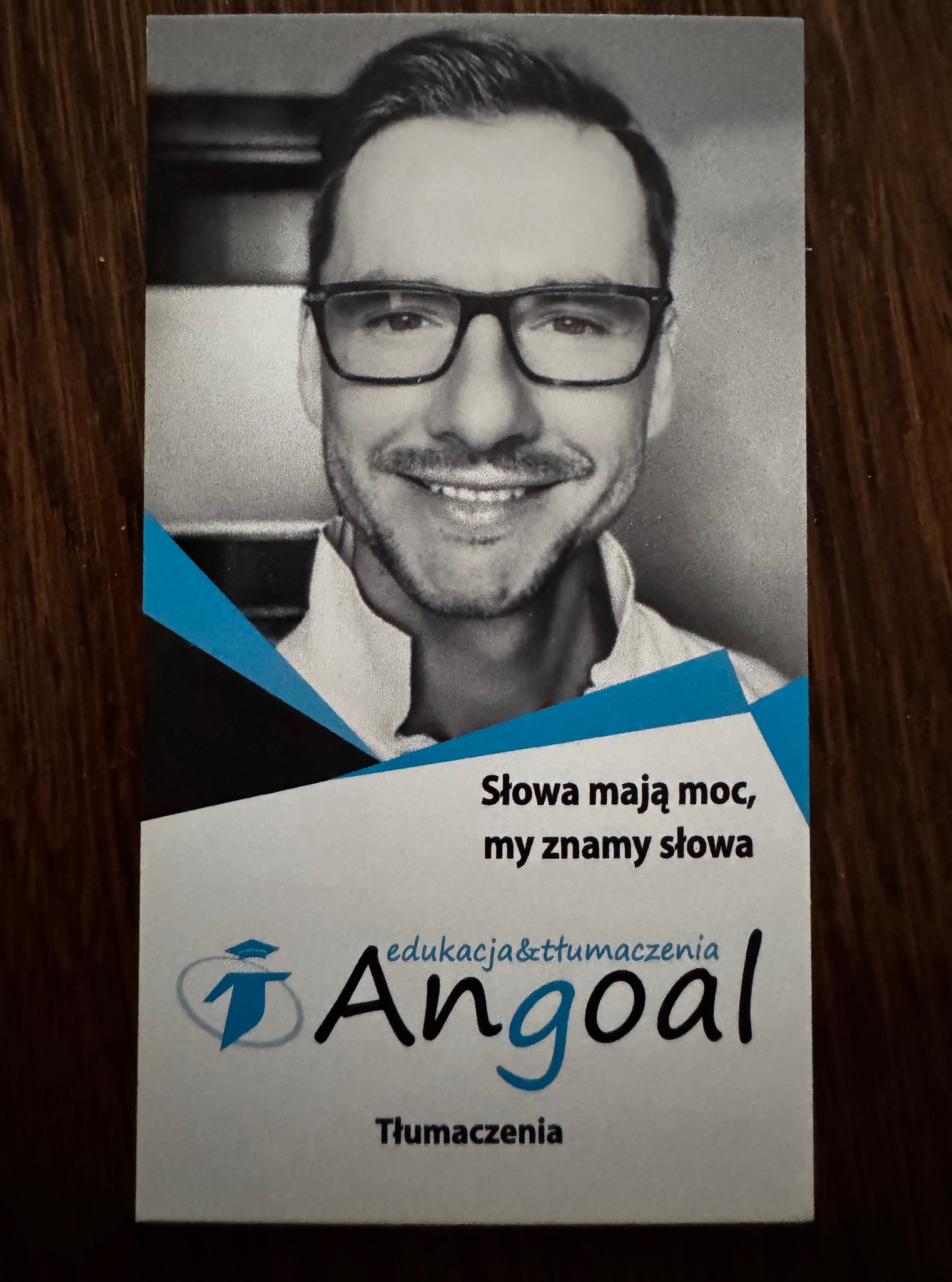 Wizytówka firmy Angoal edukacja&tłumaczenia z uśmiechniętym mężczyzną w okularach. Hasło: Słowa mają moc, my znamy słowa. Profesjonalny wizerunek.