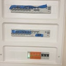 Elektro-aqbud - Biała skrzynka rozdzielcza z zabezpieczeniami, częściowo zaklejona niebieską taśmą. Widoczne bezpieczniki i diody sygnalizacyjne. Ujęcie z dołu, lekko z boku.