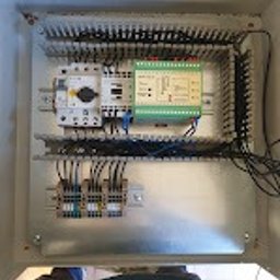 Elektro-aqbud - Wnętrze szafy sterowniczej z widocznymi elementami: przekaźniki, bezpieczniki, listwy zaciskowe i okablowanie. Ujęcie z góry, widoczne fragmenty osoby w dolnej części kadru.