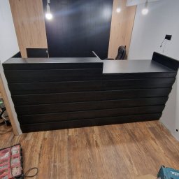 ŚWIAT MEBLI - Czarna lada recepcyjna z poziomymi panelami w Sosnowcu, na drewnianej podłodze. Widoczne narzędzia i elementy wykończeniowe, surowe oświetlenie.