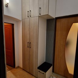 ŚWIAT MEBLI - Funkcjonalna szafa wnękowa z siedziskiem w przedpokoju, wykończona w jasnym odcieniu drewna. Minimalistyczne uchwyty, pionowe panele dekoracyjne i domowe obuwie w tle.