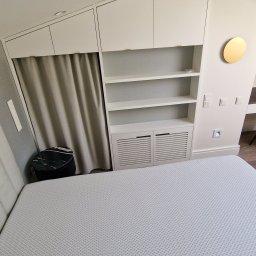 ŚWIAT MEBLI - Biała szafa wnękowa z półkami i zasłoną w pokoju na poddaszu, widok z perspektywy łóżka. Minimalistyczny design, złoty akcent dekoracyjny na ścianie.