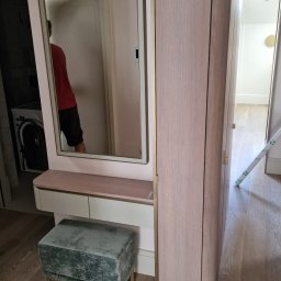 ŚWIAT MEBLI - Nowoczesna szafa z lustrem i toaletką w jasnej tonacji, uzupełniona aksamitnym pufem na złotych nóżkach. Widoczne drewniane podłogi i fragment wnętrza mieszkania.