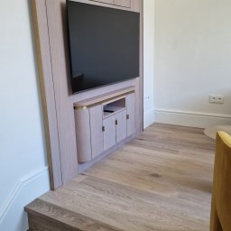 ŚWIAT MEBLI - Drewniana zabudowa TV na ścianie z szafkami i półkami, dopasowana do schodów. Minimalistyczny design, jasne drewno, złote akcenty. Nowoczesne wnętrze.