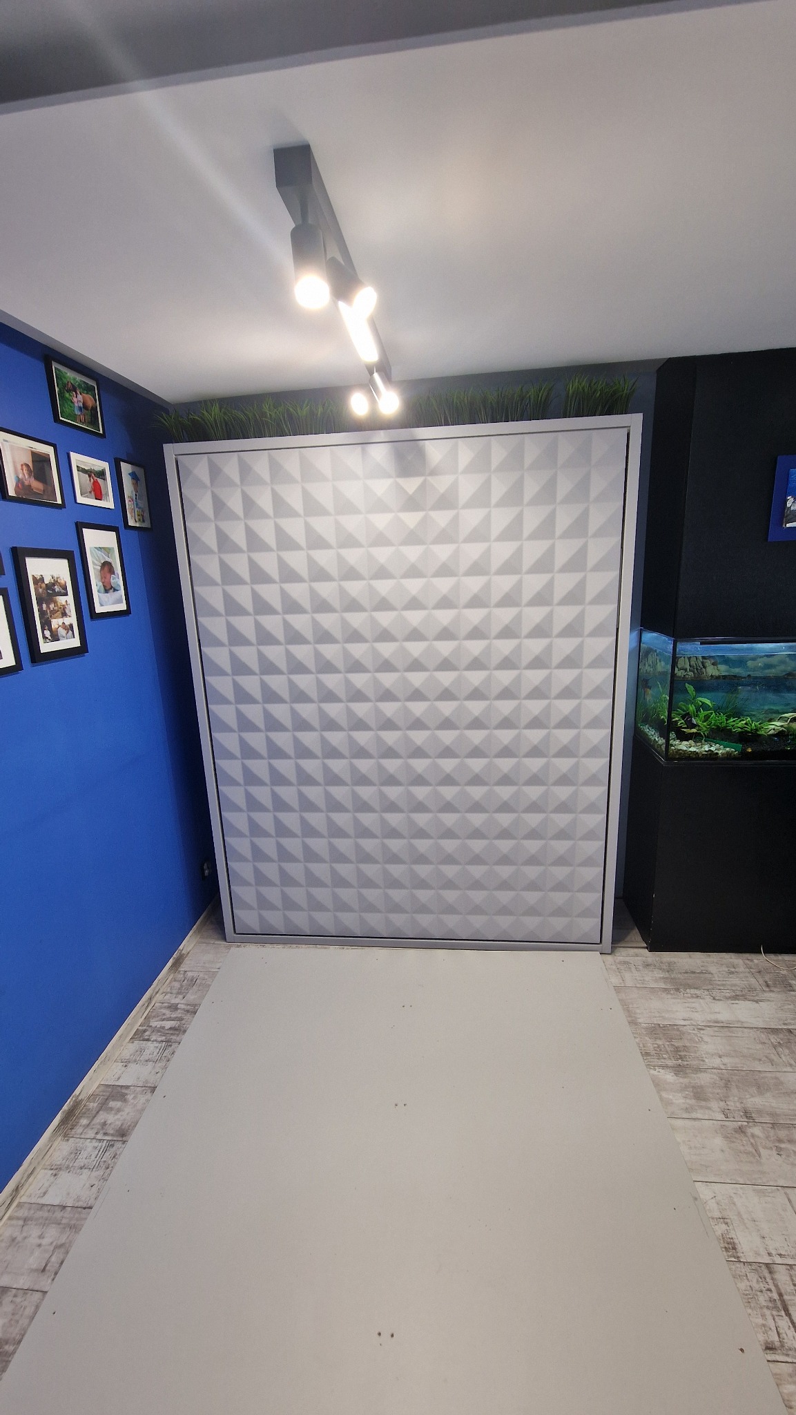 Nowoczesna szafa wnękowa z geometrycznym wzorem 3D na froncie, wkomponowana w niebieską ścianę z ramkami i sąsiadująca z akwarium. Oświetlenie sufitowe.