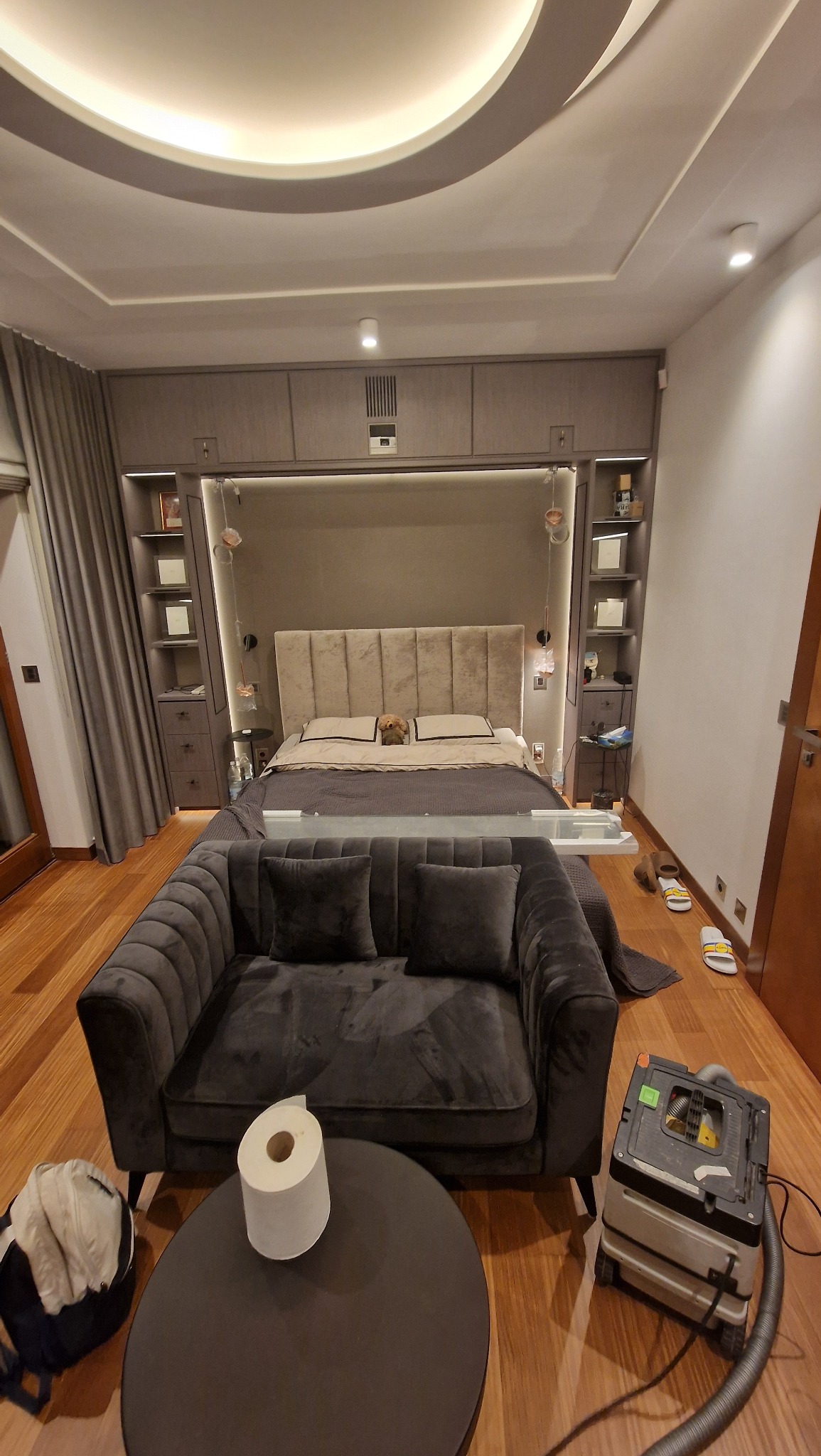 Szara szafa wnękowa z łóżkiem i tapicerowanym zagłówkiem. Wnętrze pokoju z sofą i stolikiem. Widoczny odkurzacz przemysłowy. Nietypowe oświetlenie sufitowe.