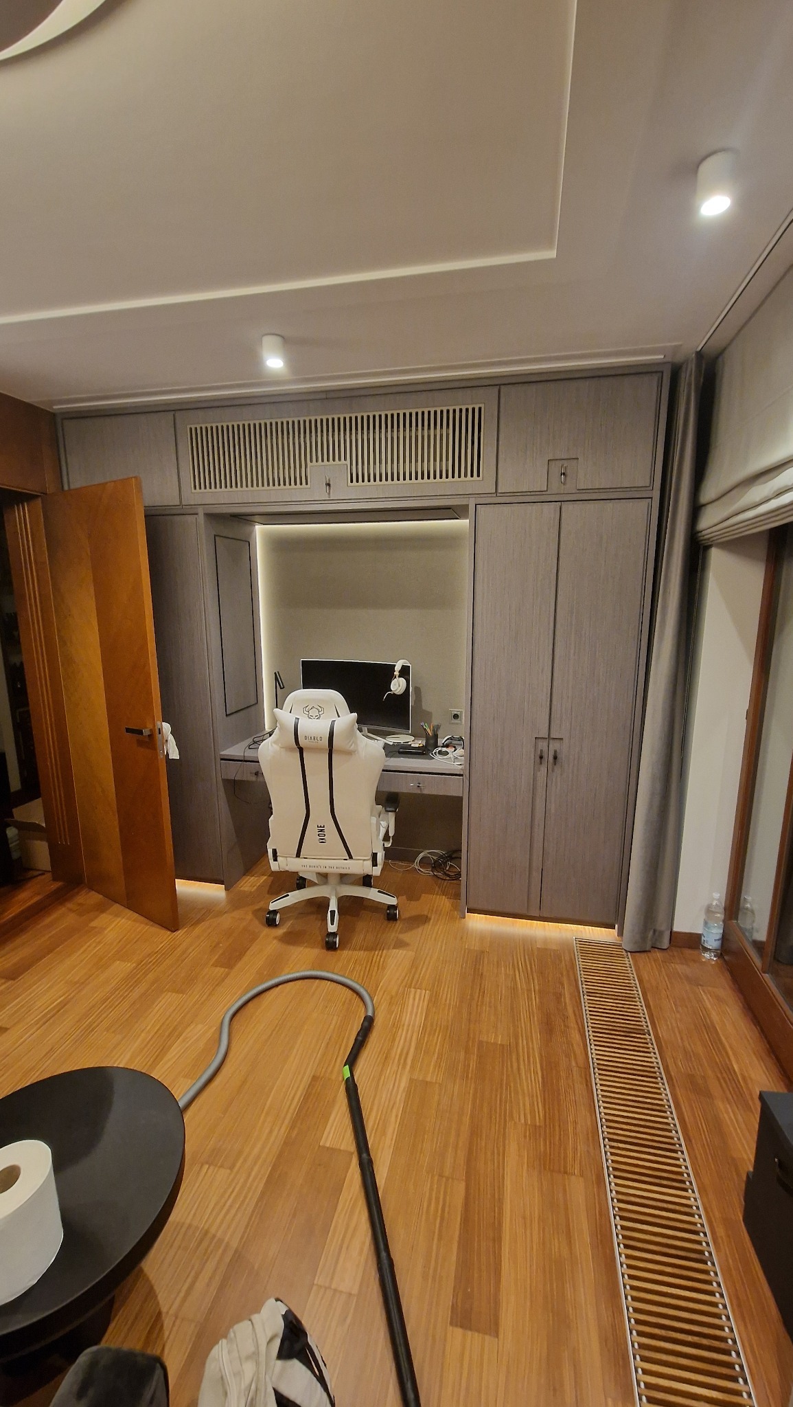 Szara szafa wnękowa z biurkiem i oświetleniem LED, wbudowana w ścianę. Na biurku stoi białe krzesło gamingowe i monitor. Widok na drewnianą podłogę.