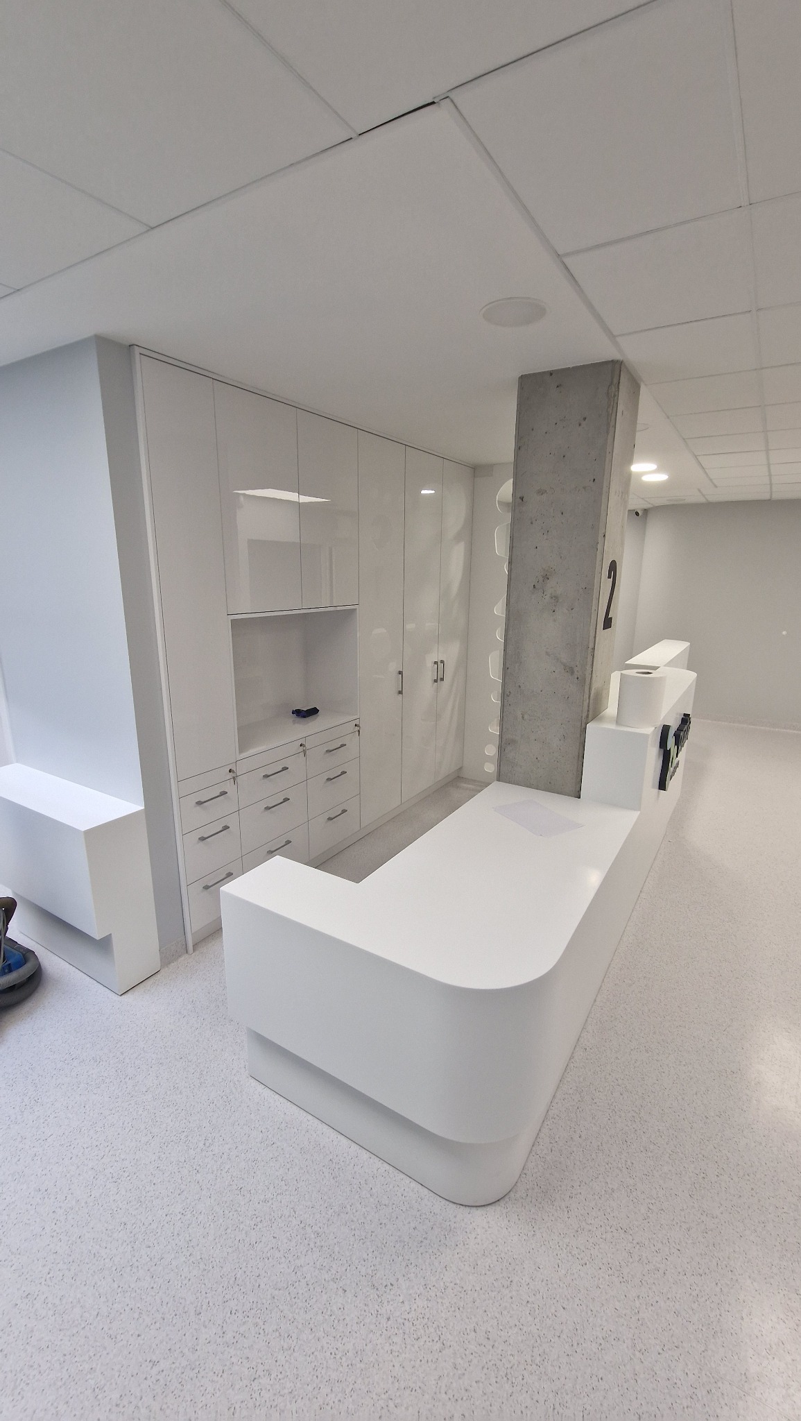 Biała lada recepcyjna z szafkami w tle, minimalistyczny design, oświetlenie sufitowe, betonowy filar z numerem '2', jasna podłoga z drobnym wzorem.