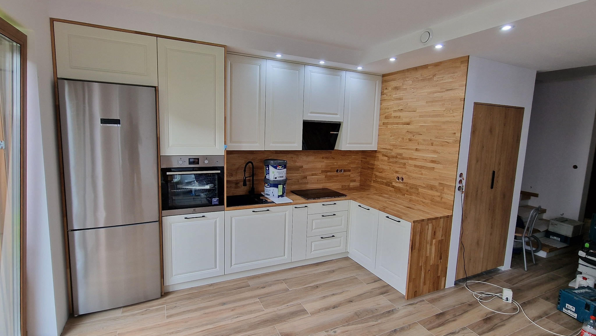 Jasna kuchnia z drewnianymi akcentami: białe szafki, drewniany panel na ścianie, stalowa lodówka. Widoczne farby i narzędzia, sugerujące etap wykończeniowy.