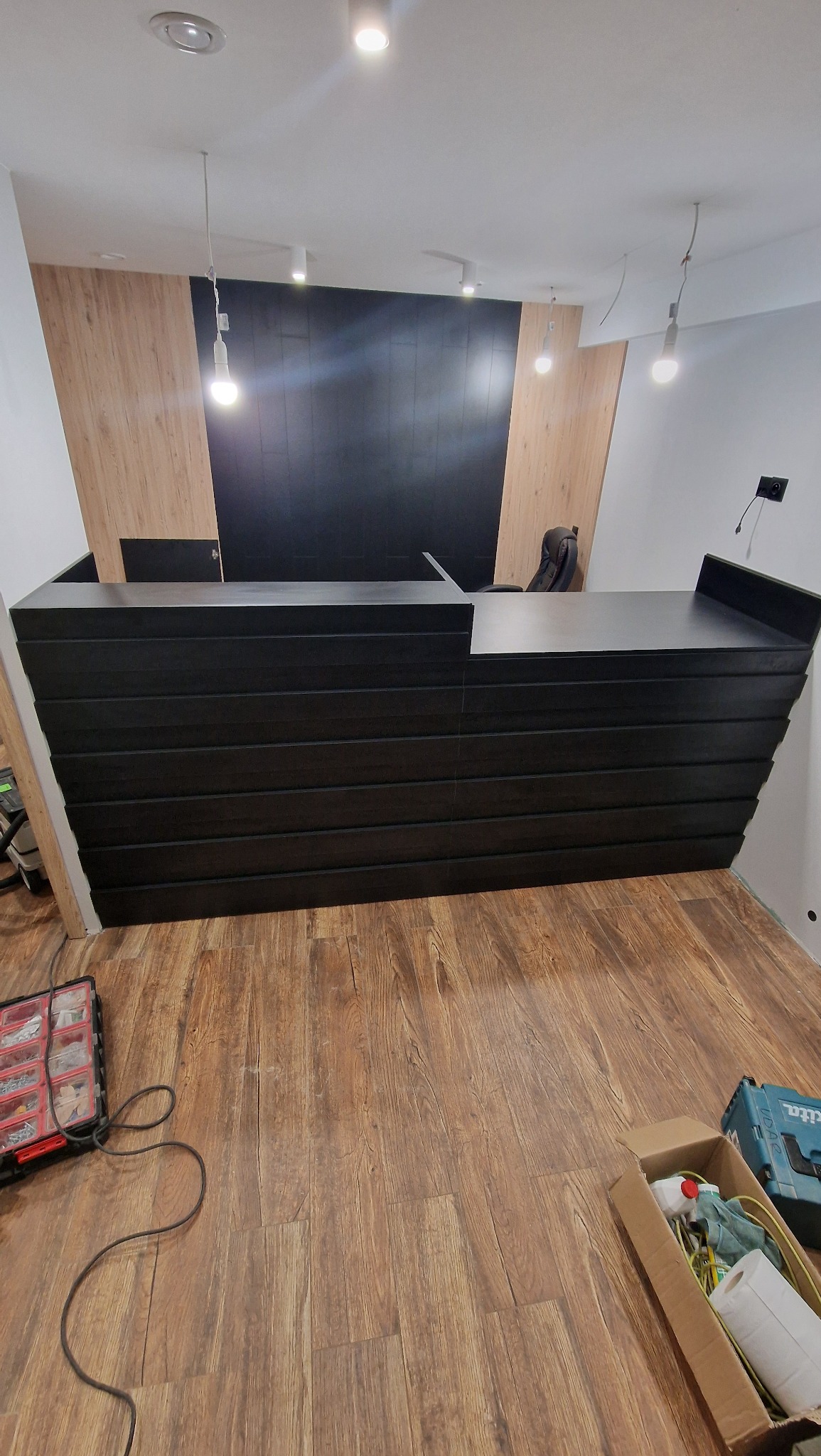 Czarna lada recepcyjna z poziomymi panelami w Sosnowcu, na drewnianej podłodze. Widoczne narzędzia i elementy wykończeniowe, surowe oświetlenie.