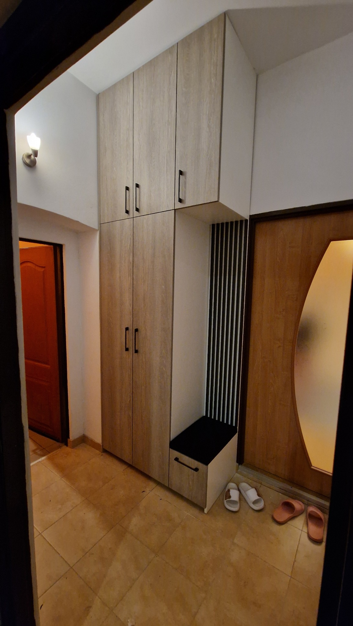 Funkcjonalna szafa wnękowa z siedziskiem w przedpokoju, wykończona w jasnym odcieniu drewna. Minimalistyczne uchwyty, pionowe panele dekoracyjne i domowe obuwie w tle.