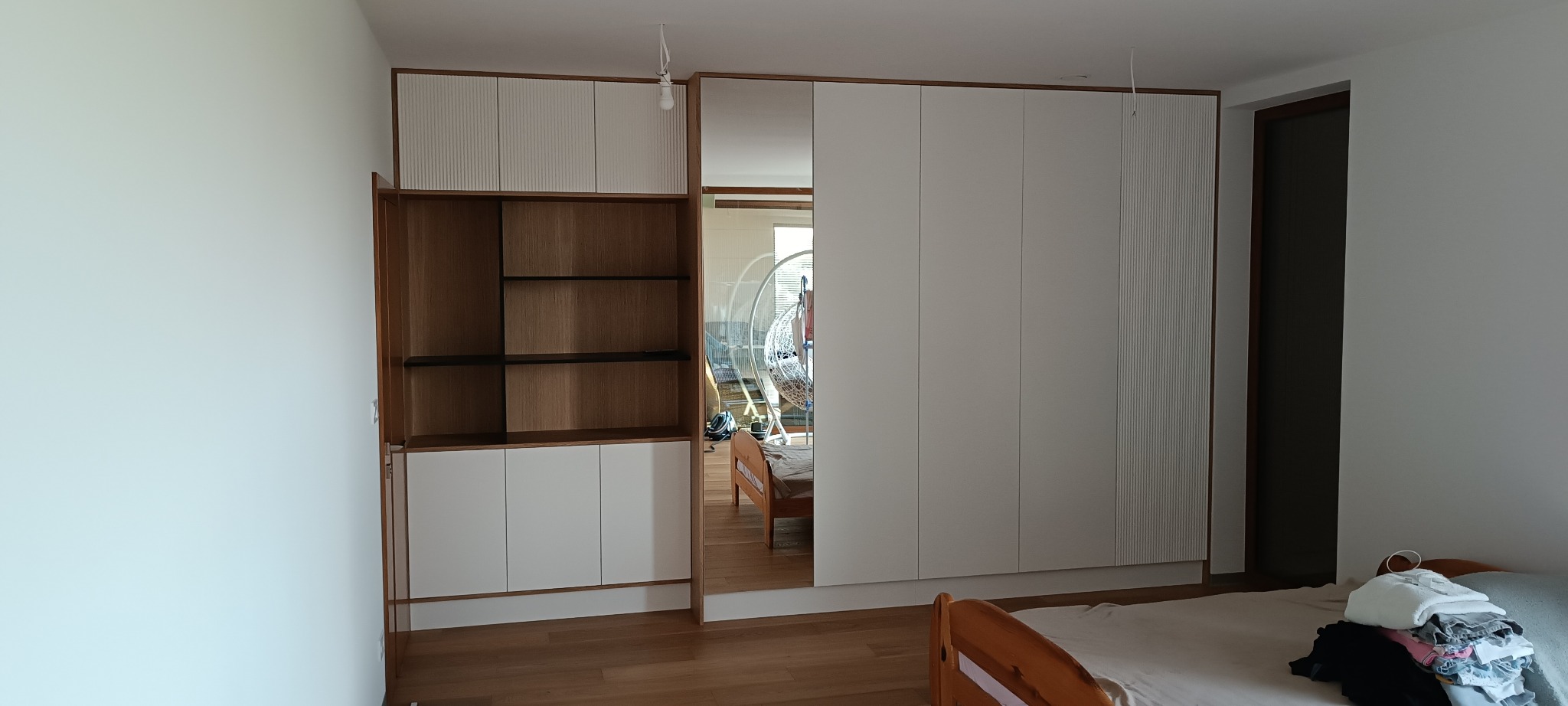 Biała szafa wnękowa z lustrem i regał z drewnianymi półkami, wkomponowane w jasne wnętrze sypialni. Widoczne detale wykończenia i odbicie pokoju w lustrze.