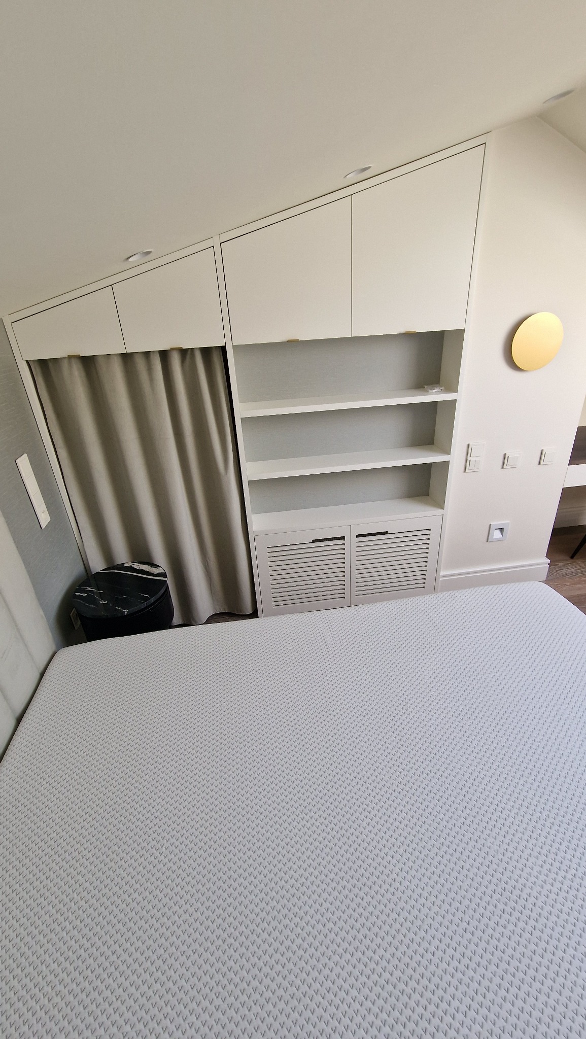 Biała szafa wnękowa z półkami i zasłoną w pokoju na poddaszu, widok z perspektywy łóżka. Minimalistyczny design, złoty akcent dekoracyjny na ścianie.