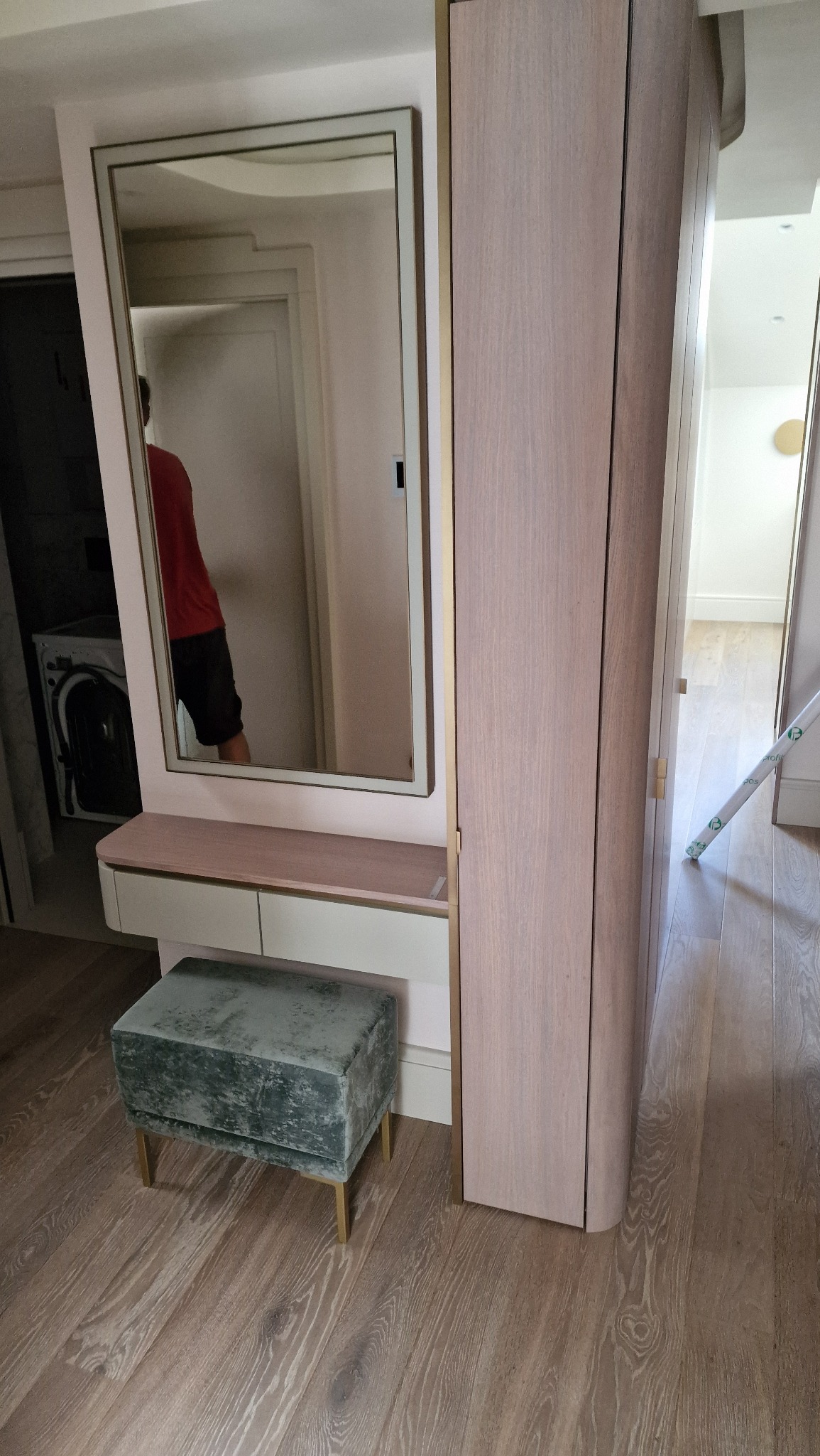 Nowoczesna szafa z lustrem i toaletką w jasnej tonacji, uzupełniona aksamitnym pufem na złotych nóżkach. Widoczne drewniane podłogi i fragment wnętrza mieszkania.
