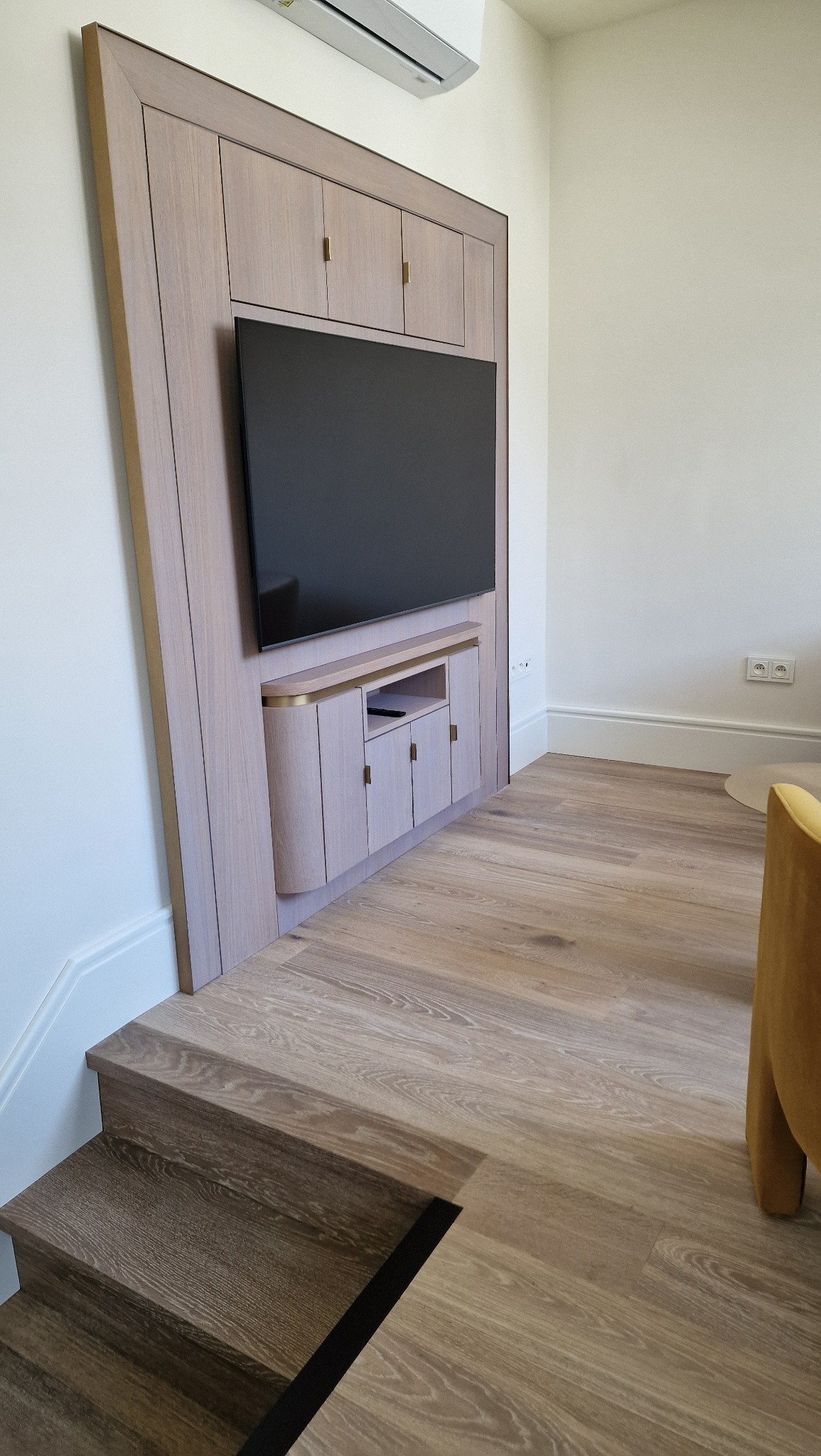 Drewniana zabudowa TV na ścianie z szafkami i półkami, dopasowana do schodów. Minimalistyczny design, jasne drewno, złote akcenty. Nowoczesne wnętrze.