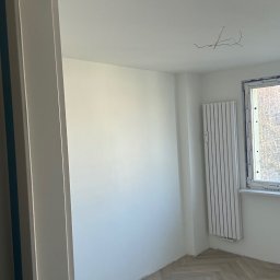 Miłosz Panek - Jasne wnętrze po remoncie: świeżo pomalowane ściany, nowy parkiet w jodełkę, biały grzejnik pod oknem. Widoczne wystające kable ze ściany.