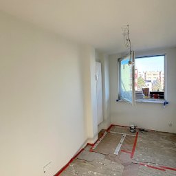 Miłosz Panek - Jasne wnętrze po malowaniu, z zabezpieczoną podłogą i otwartym oknem z widokiem na budynki. Świeżo pomalowane ściany, widoczne narzędzia malarskie.