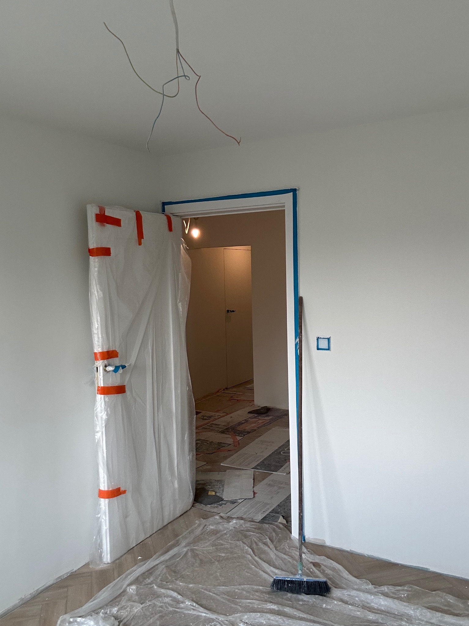 Białe ściany po malowaniu, zabezpieczone drzwi folią i taśmą, widoczne kable zwisające z sufitu, podłoga częściowo zakryta folią ochronną, pomieszczenie w trakcie prac wykończeniowych.