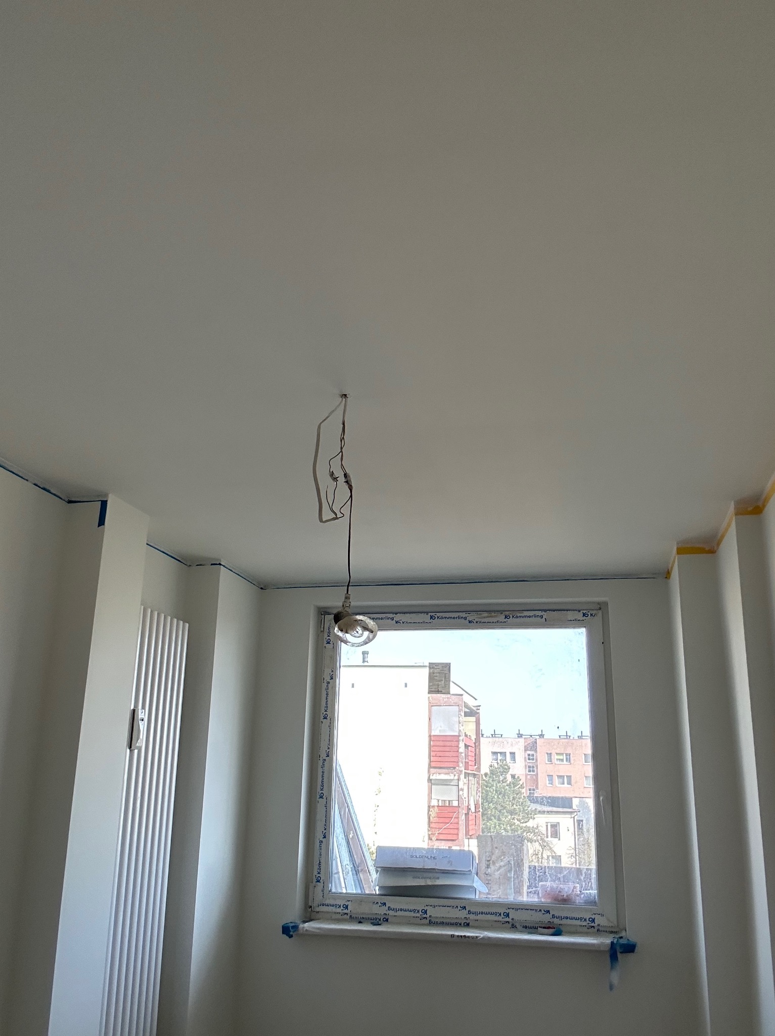 Białe wnętrze pokoju po malowaniu, widoczne taśmy malarskie przy suficie i oknach. Lampa zwisa z sufitu. Widok na budynki przez okno.
