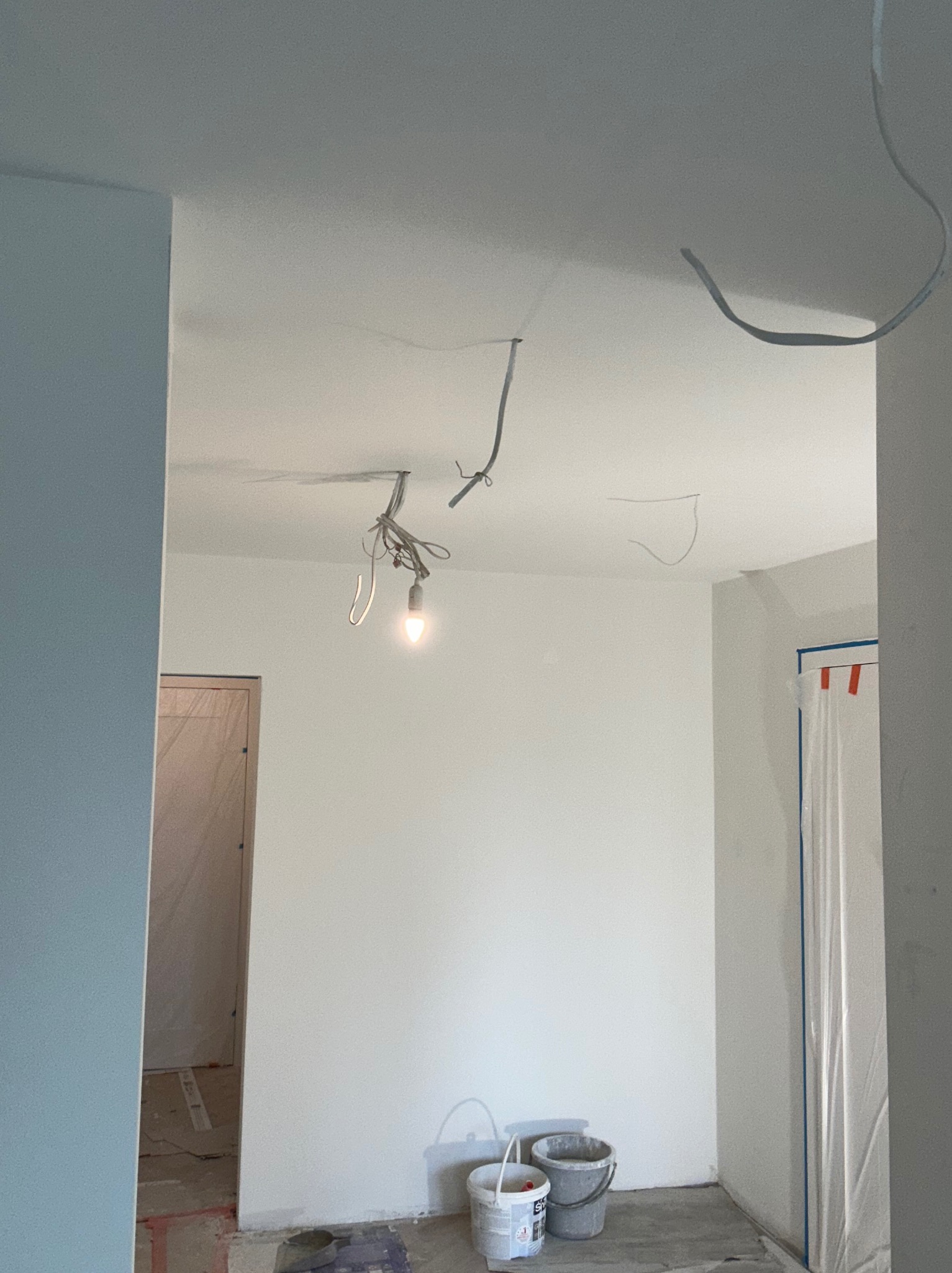 Wnętrze w trakcie prac: białe ściany, wiszące kable, żarówka, wiaderka z farbą na podłodze. Drzwi zabezpieczone folią malarską. Widok z perspektywy podłogi.