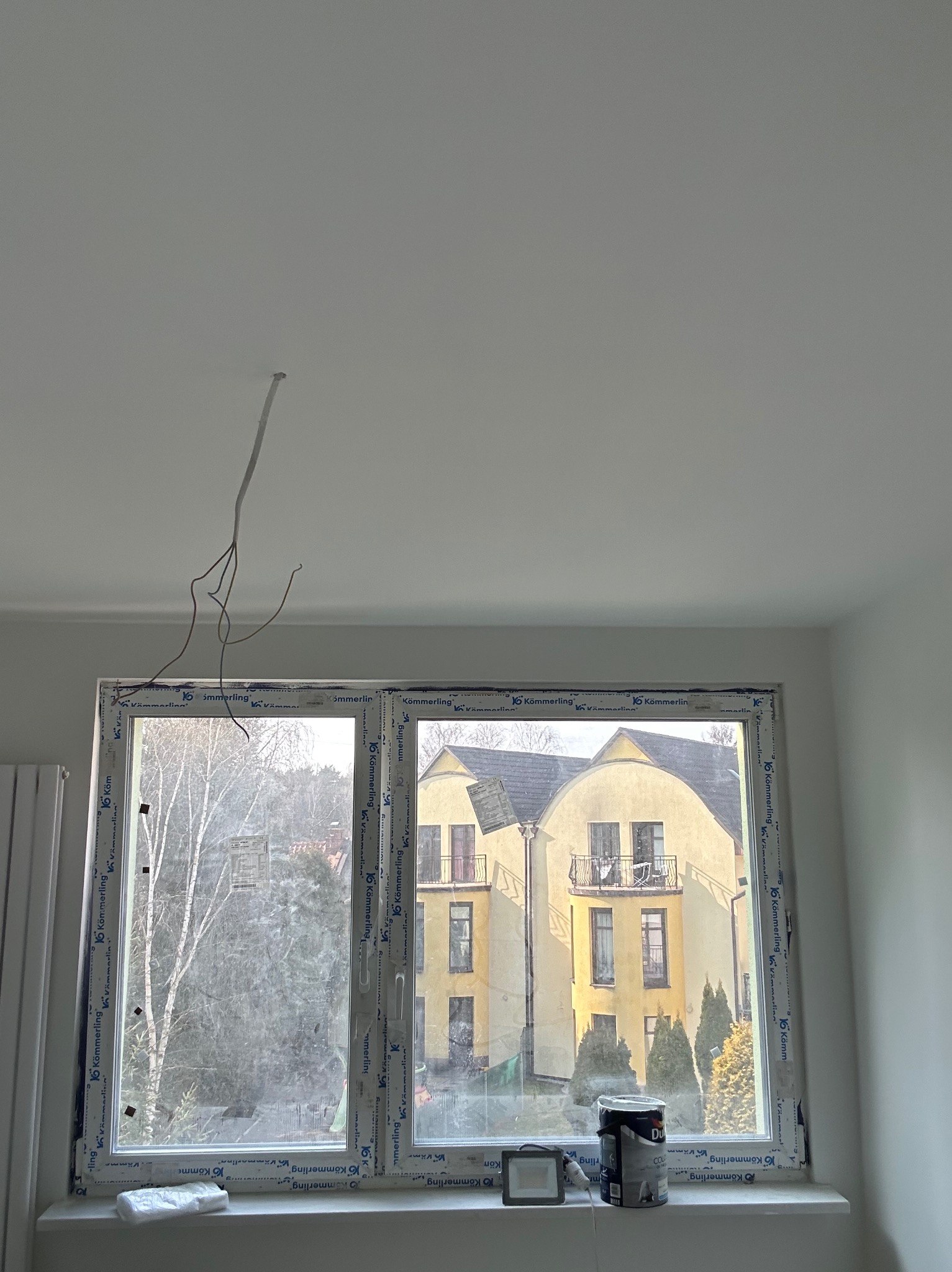 Białe okno PCV z widokiem na budynek. Na parapecie puszka farby Dulux i lampa. Z sufitu zwisają kable. Świeżo pomalowane ściany i sufit. Remont w toku.