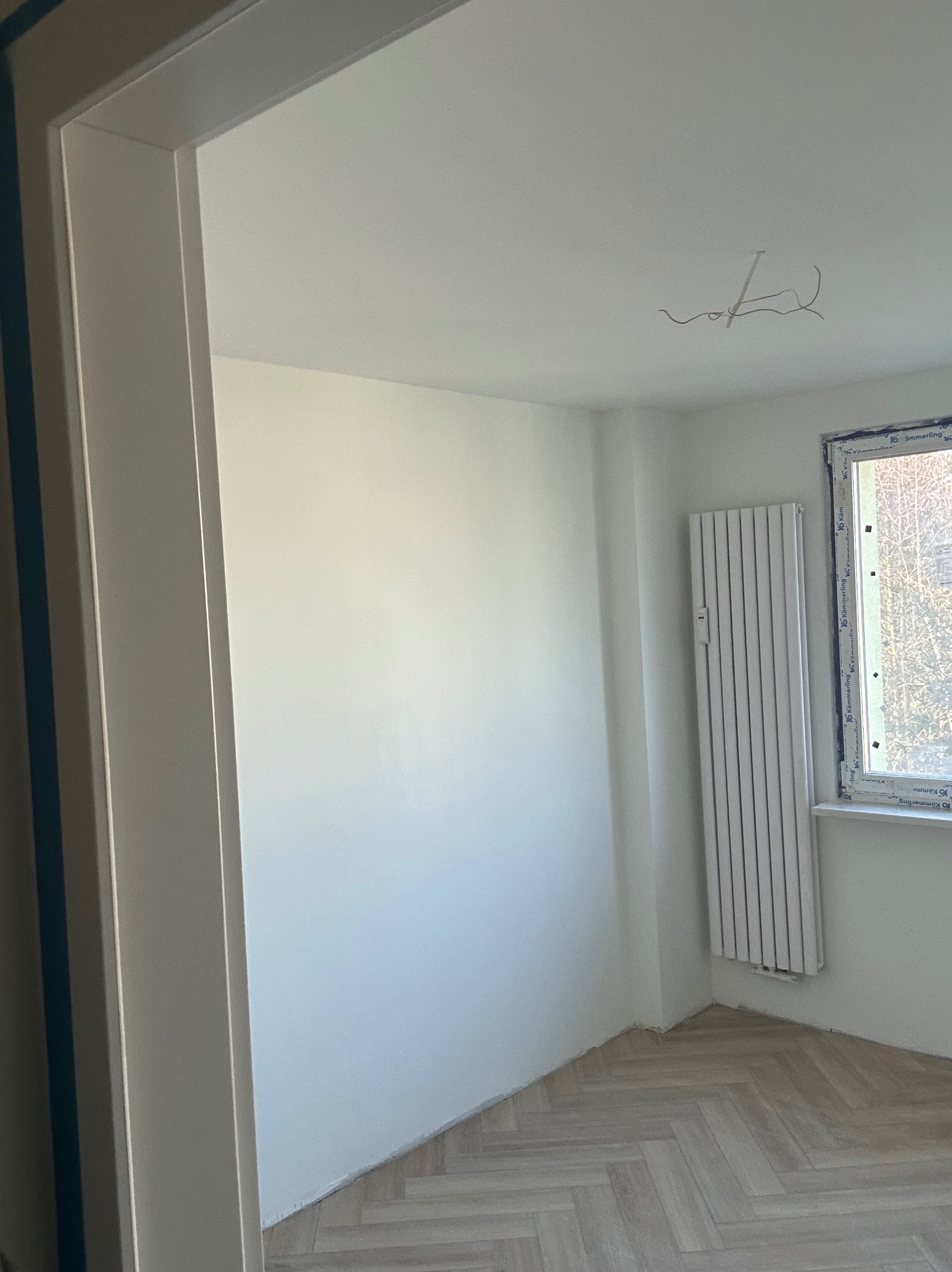 Jasne wnętrze po remoncie: świeżo pomalowane ściany, nowy parkiet w jodełkę, biały grzejnik pod oknem. Widoczne wystające kable ze ściany.