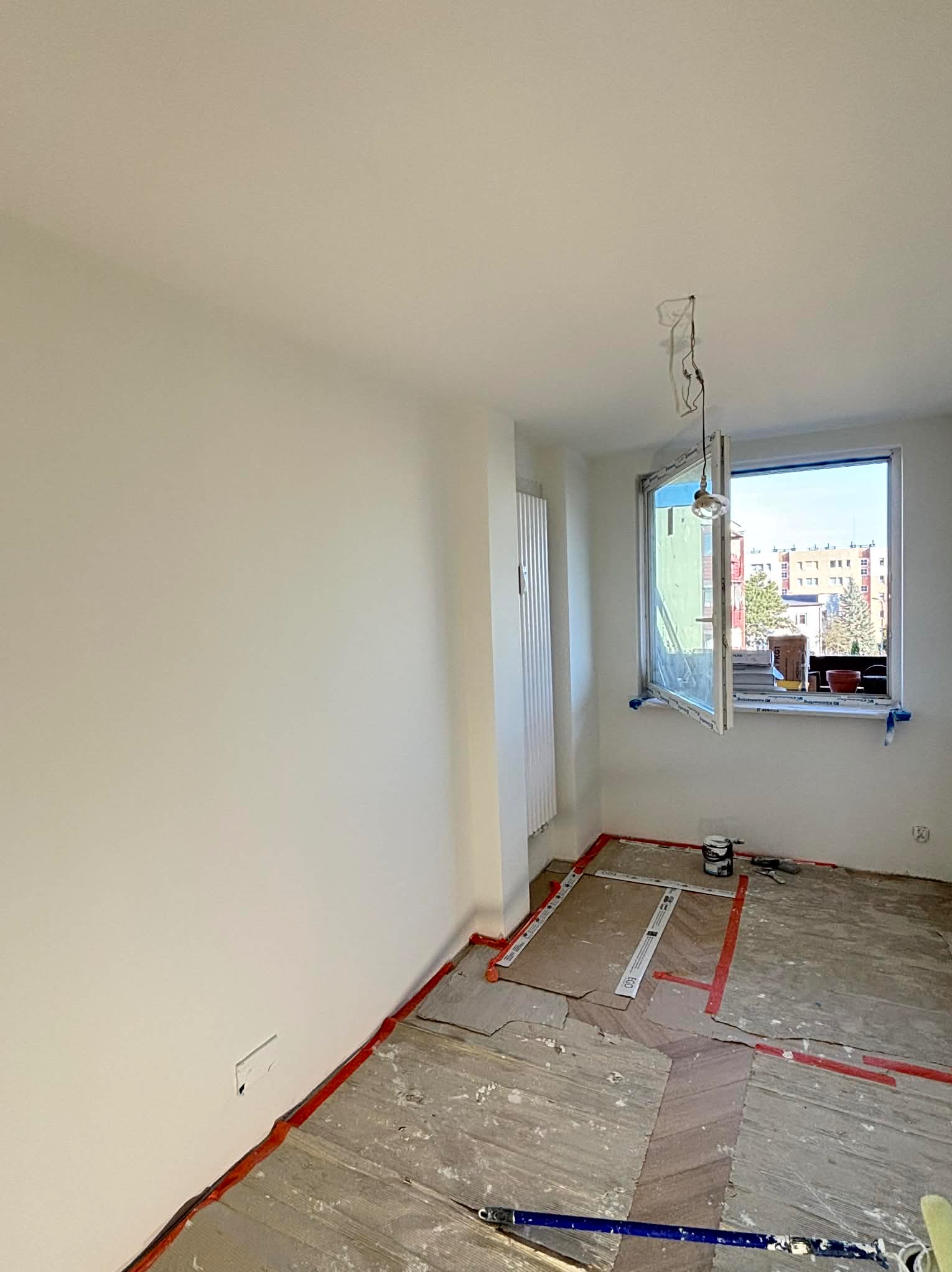 Jasne wnętrze po malowaniu, z zabezpieczoną podłogą i otwartym oknem z widokiem na budynki. Świeżo pomalowane ściany, widoczne narzędzia malarskie.