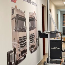WallKing Łukasz Żugaj - WallPen drukuje na ścianie w biurze grafiki dwóch ciężarówek Kigar Transport Logistyka. Drukarka na szynach, w tle drzwi i korytarz.