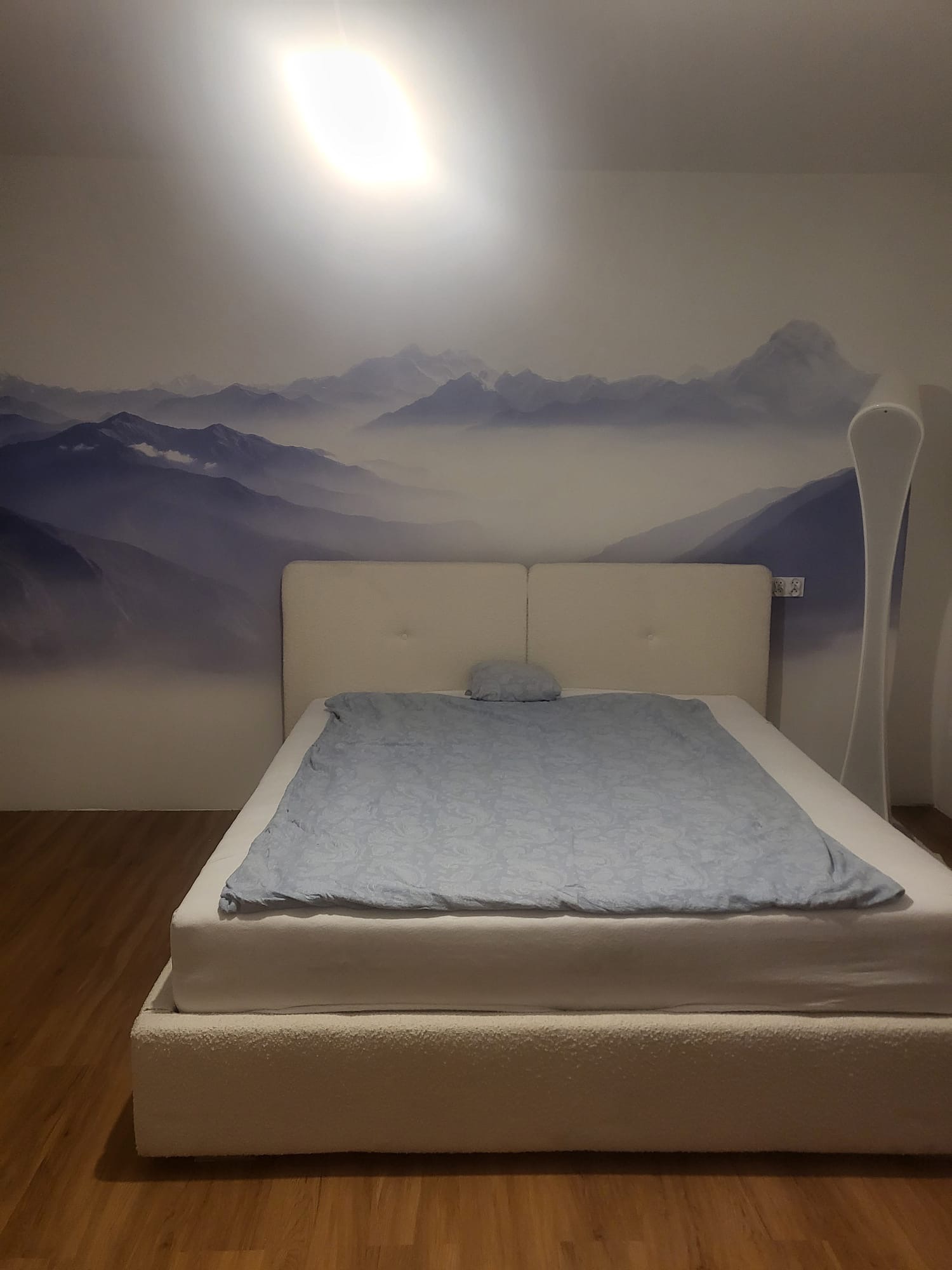 Sypialnia z łóżkiem i fototapetą górskiego krajobrazu. Białe łóżko z jasnoniebieską pościelą. Na ścianie fototapeta z motywem gór w odcieniach szarości i błękitu.