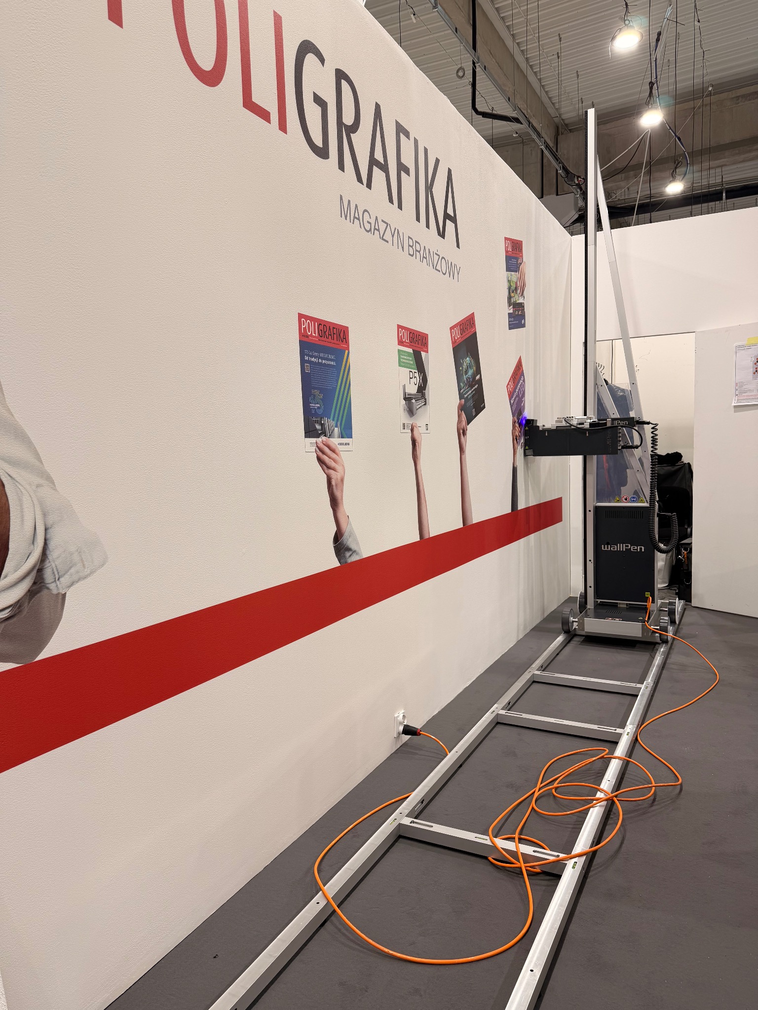 WallPen drukuje grafikę na ścianie. Widoczny magazyn branżowy 'Poligrafika' i czerwony pas dekoracyjny. Urządzenie na szynach, pomarańczowy kabel zasilający.